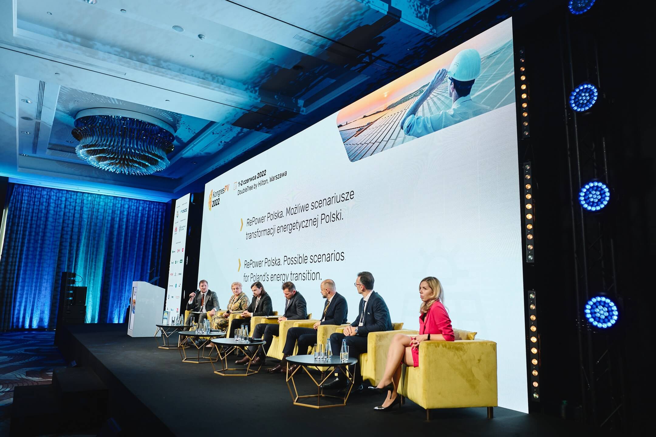 Spotkanie branży PV z udziałem pracowników Projekt Solartechnik. Przygaszone światła w sali konferencyjnej. Na środku scena, na której zasiada na musztardowych fotelach siedmioro ekspertów. Za nimi wielki ekran na którym na białym tle wyświetlana jest prezentacja dotycząca OZE.