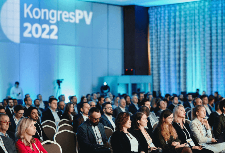 Sala konferencyjna wypełniona bardzo duża ilością osób będących uczestnikami spotkania branży PV. Na ścianie za nimi wyświetlany jest napis kongres PV 2022. Światło jest przygaszone, wszyscy uczestnicy patrzą przed siebie na prezentowane treści związane z odnawialnymi źródłami energii.