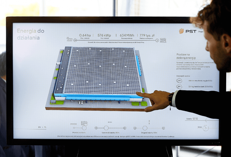 Olbrzymi telewizor, na którym wyświetlana jest wizualizacja możliwości wsparcia biznesu i czerpania zysku z umieszczenia fotowoltaiki na dachu firmy. Pracownik Projekt Solartechnik omawia możliwości współpracy wskazując palcem interesujące dane.